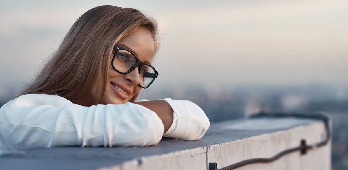 Porträt einer entspannten jungen Geschäftsfrau mit Brille, die in der Abenddämmerung auf dem Balkon steht und die Stadt überblickt. 